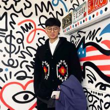 A person in a black knitted cardigan, holding a blue jacket and iphone. They're standing in front of a white wall with swirly shapes and a heart with an eye in it.