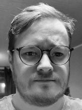 A black and white image of Dean Chalmers, looking to the camera and wearing glasses.