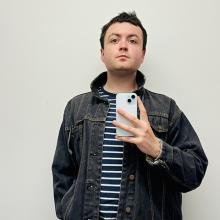 Ewan Pearce takes a photo of themselves in a mirror. They are wearing a blue striped top and washed out black denim jacket.