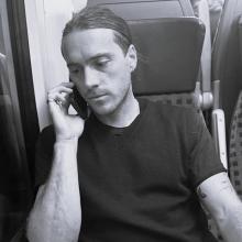 A black and white photo of Ian sitting on a train, holding a phone to his ear.