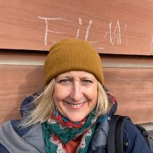 Tracey stands outside the Main Building ECA, under a chalk sign that reads 'Film'. They wear a waterproof coat, scarf and mustard beanie hat.