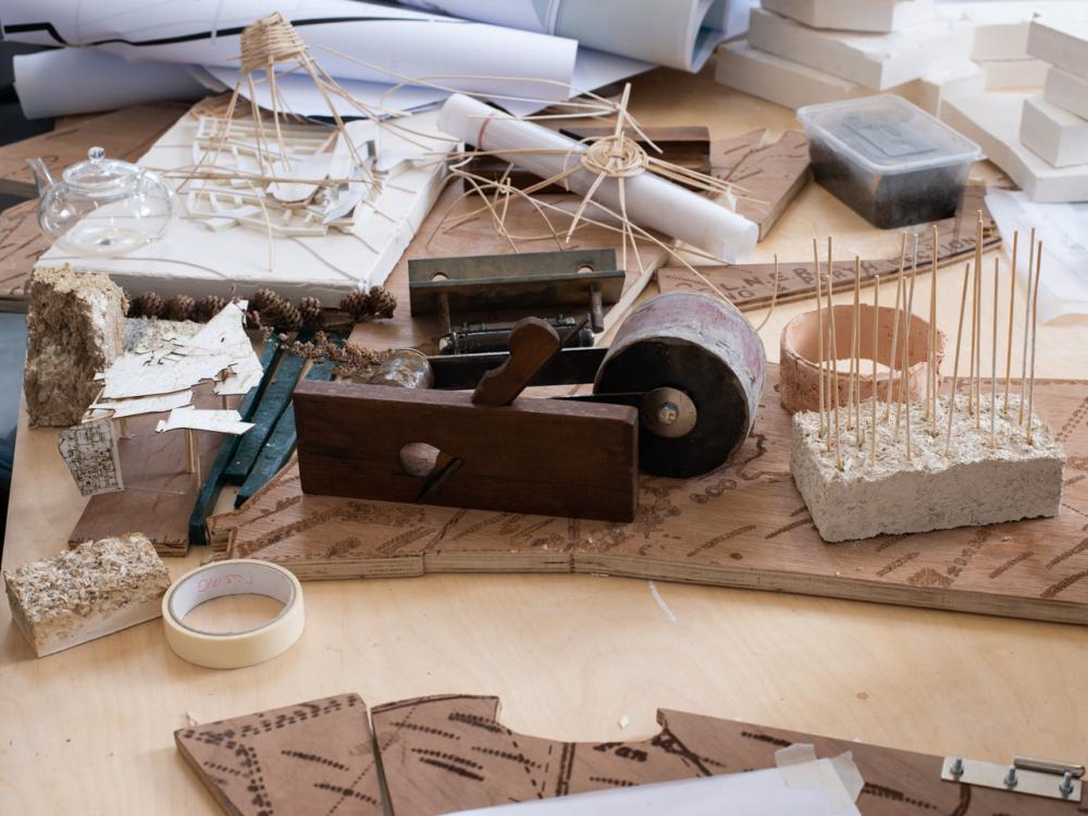 A collection of materials and tools on a desk, including rolled up paper, pieces of wood, tape, cutting tools and a plastic box
