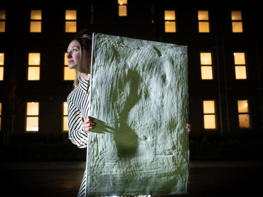Student holding their artwork outside an apartment block at night with lights in the windows.