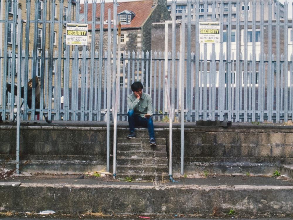 A person sits outside, at the top of concrete steps in front of a metal fence