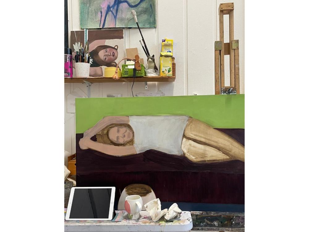 A painting of a reclining woman, on an easel. There's a shelf of paints and brushes on the wall behind