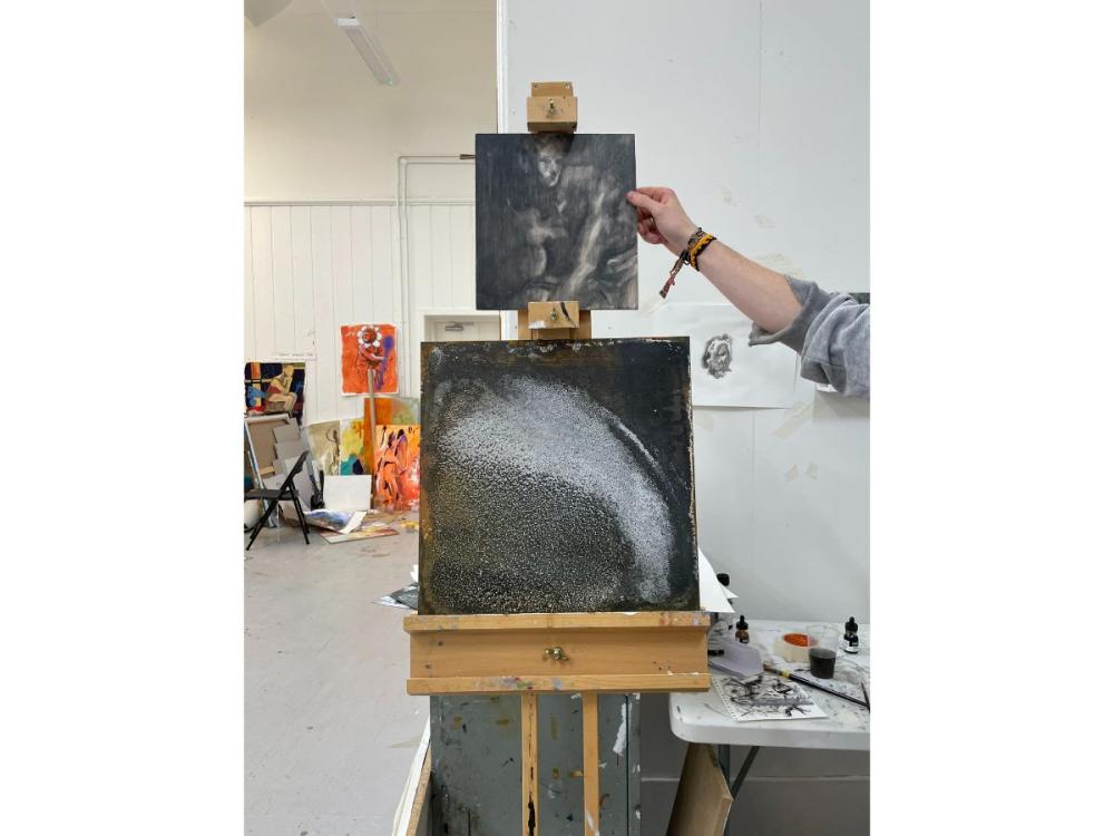 Two works on an easel, both in shades of black, grey and white. The bottom image is abstract, a black background with a swirl of white across it; the top work is a shadowy image of a figure leaning forward.