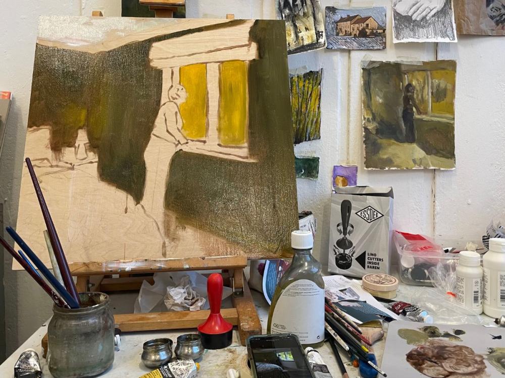 A work in progress painting of a woman looking out of a window. On the wall behind are more paintings and sketches; on the table alongside is a jar with paintbrushes in, and a selection of other materials