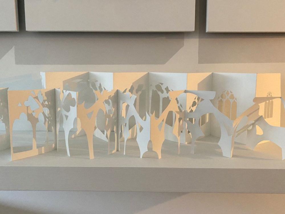 A series of beautifully-lit, interlinked cut-outs in white card. The back row is suggestive of church windows, the front row more abstract