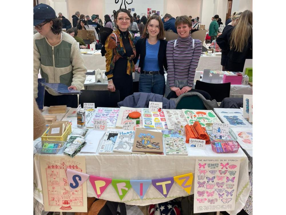 The three designers standing behind their stall at Bookmarks. They're smiling at the camera; their work on the stall includes colourful prints and zines