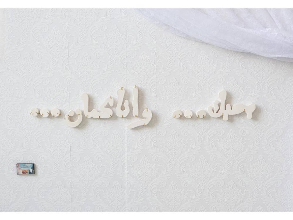 Arabic letters in white plaster, raised up from the white background on which they sit. To the top right is a piece of white fine netting or mesh material, on the bottom left is a cassette tape in a box, titled 'Spoken Arabic'.