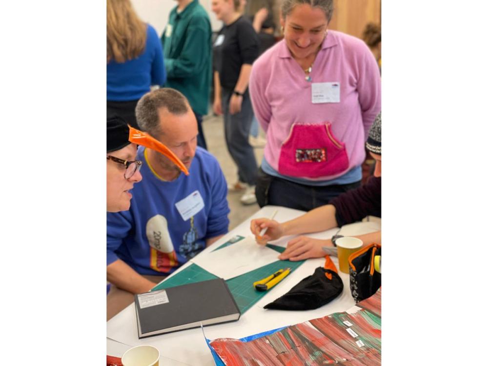 Several illustrators gathered together at a table, chatting. One is holding a pen and about to draw on a piece of plain white paper