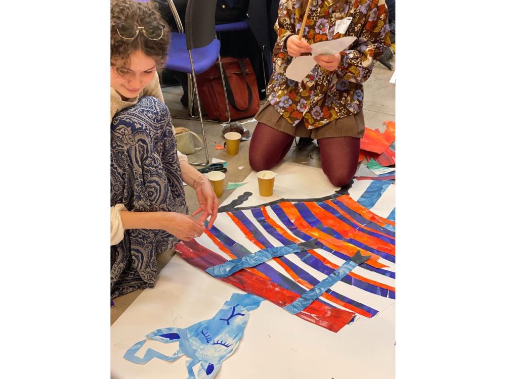 Two illustrators sitting on the floor, working on a collage of a blue animal, sleeping in a bed. 