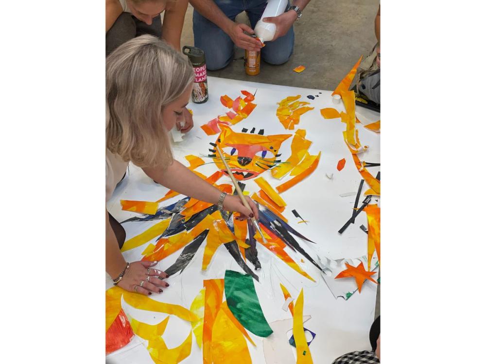 An illustrator sitting on the floor, painting onto a colourful collage of a cat