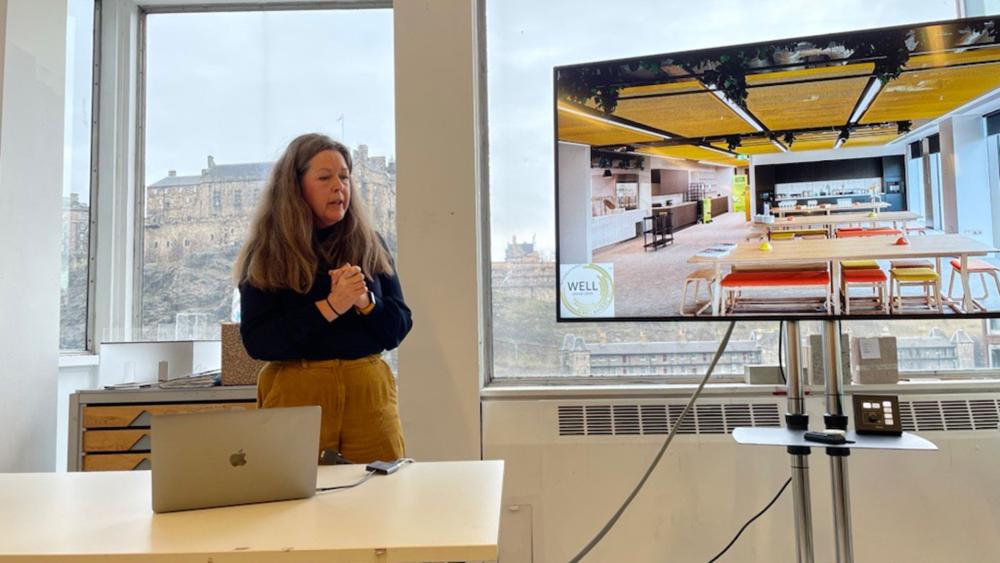 A person with long hair stands next to a big tv screen with a picture of an interior office space on it.