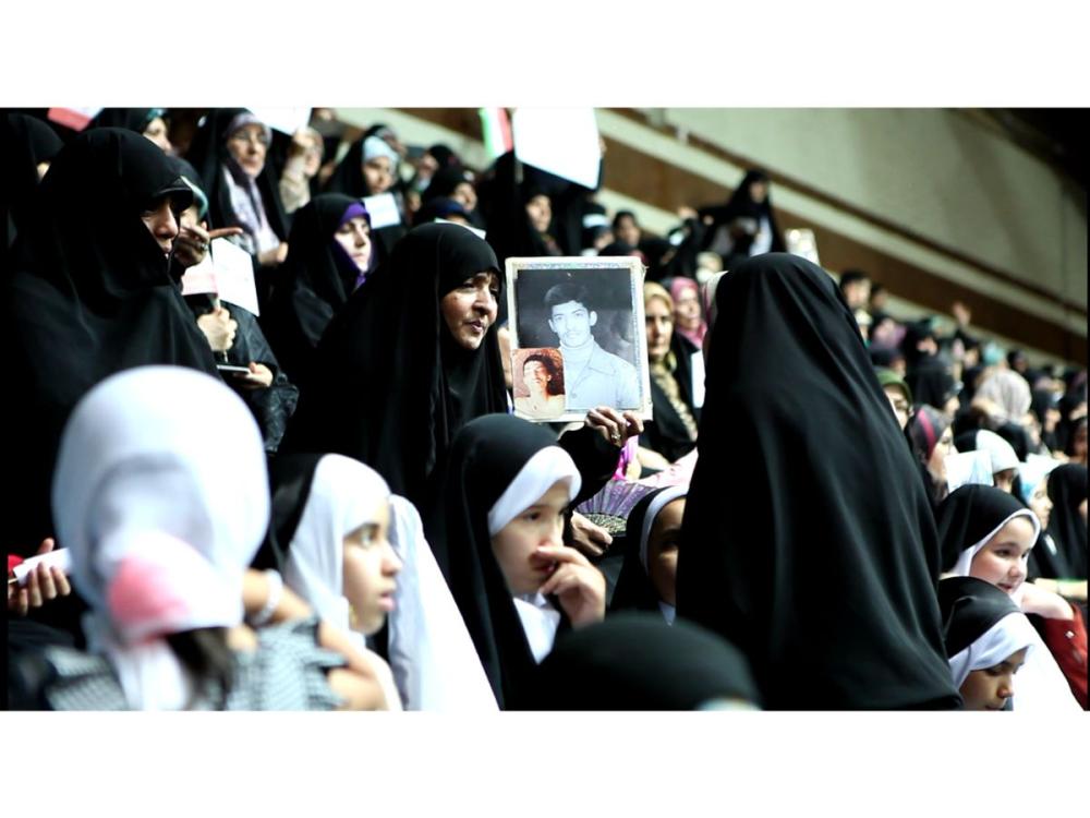 A crowd of women and girls, wearing hijab. They look to be on raked seats or platforms - perhaps in a stadium. In the centre, a woman holds up two head-and-shoulders photographs of a young man.
