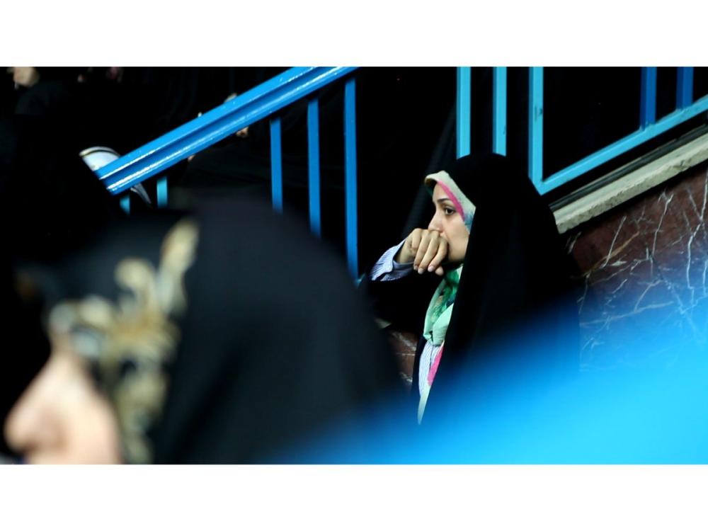 A young woman wearing hijab leans against a wall, perhaps the entrance steps to a stadium. She looks worried, with the back of her hand over her mouth.