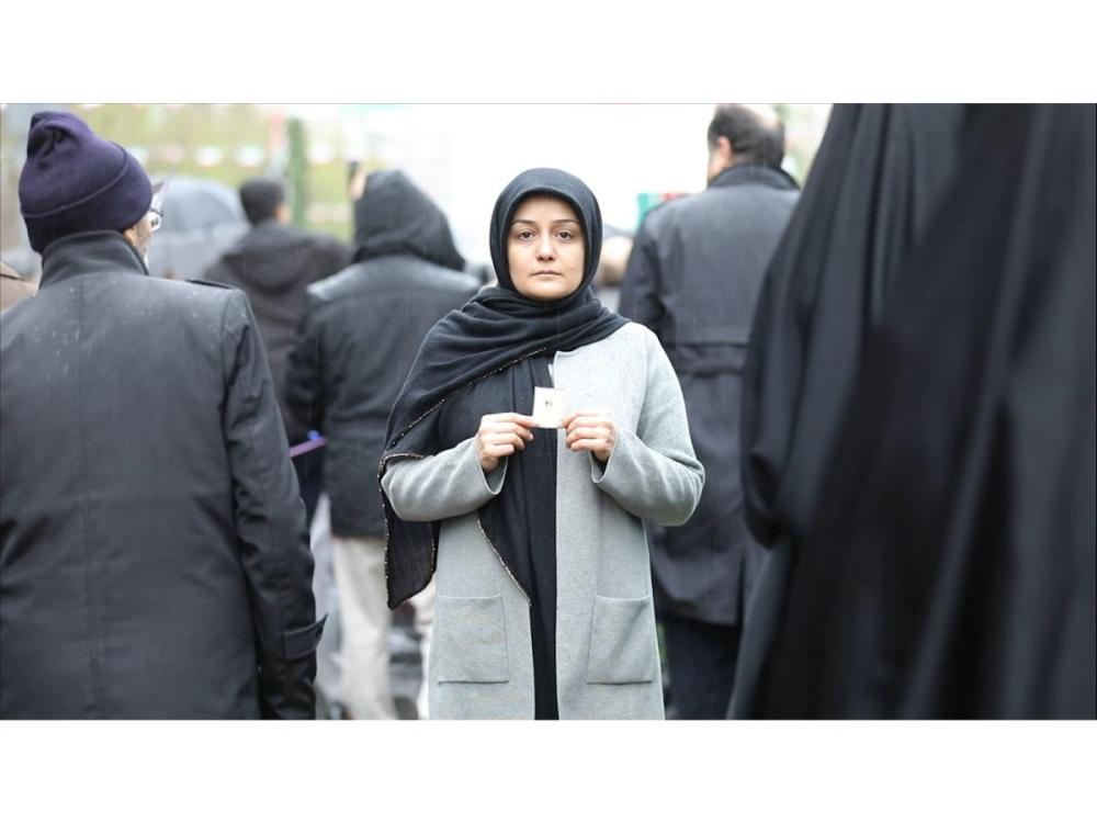 A woman in black hijab and a grey coat looks straight at the camera with a solemn expression, holding a small square of paper with both hands. She's in a crowd - the other people, all men, wearing black, are walking away from their camera - we see only their backs.