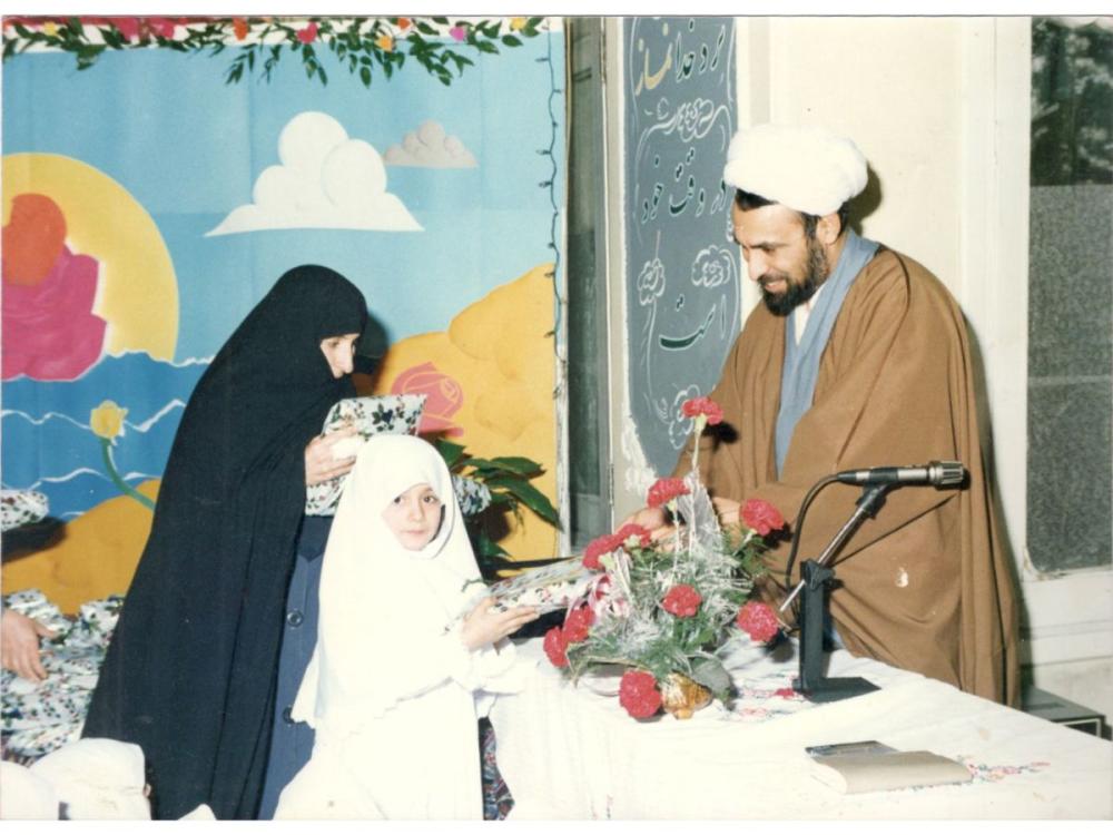 A childhood photograph. A woman and child stand on one side of a table. The child is wearing white hijab, the woman, a teacher, wearing a black chador (cloak). On the other side of the table stands a smiling man - a cleric - wearing an aba (rob) and amameh (turban). The table has flowers on it; behind them is a colourful mural or painting showing sea, sand, sky and sun.