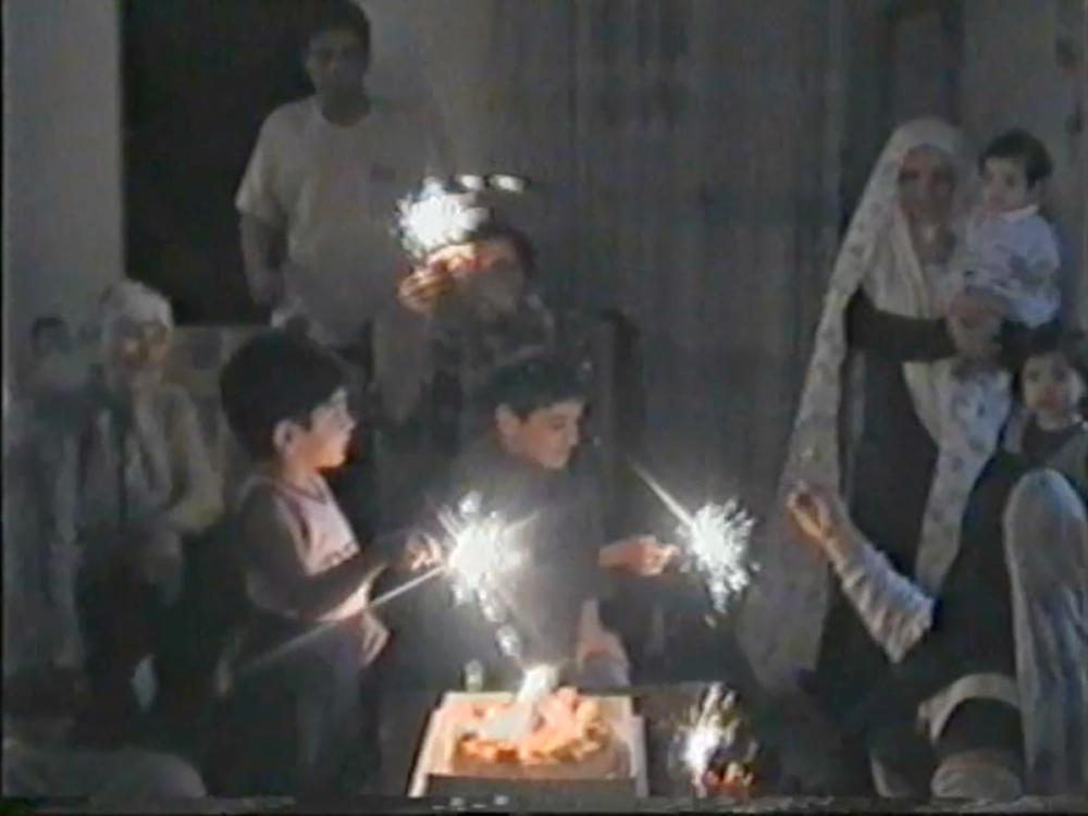 A dark room - children standing around a cake hold sparklers; Adults stand behind them, one holding a smaller child.