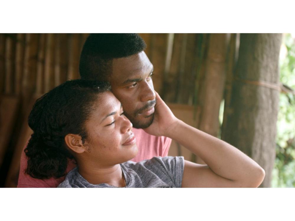 A man and a woman stand together. Their heads rest against one another; her eyes are closed, he looks straight ahead. She is reaching up over her shoulder, and cupping his chin in her hand.