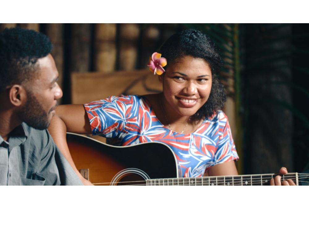 A smiling woman with a flower in her hair plays the guitar; a man looks on.