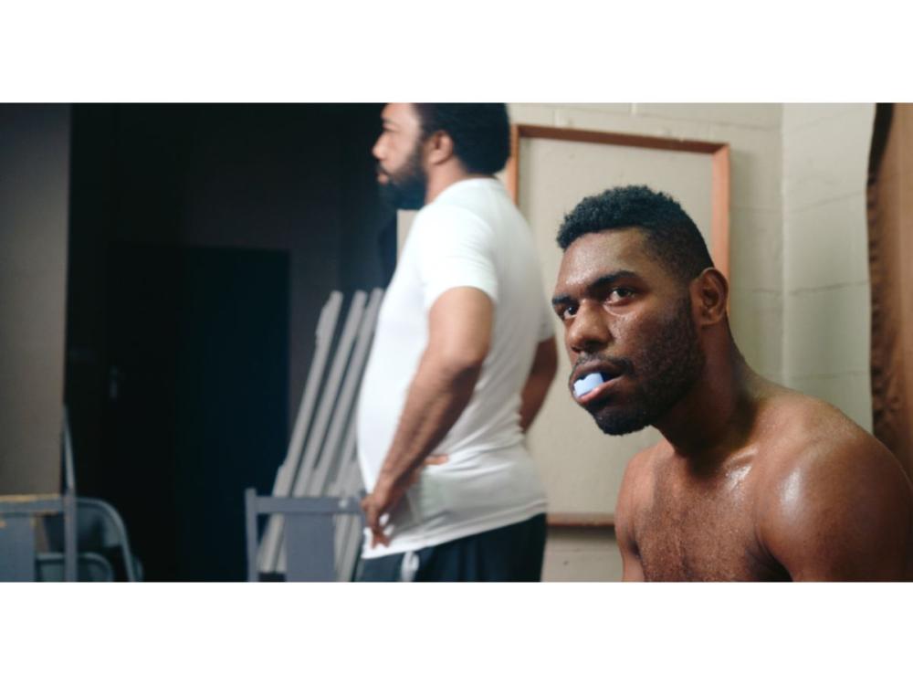 A man with a bare chest and a mouthguard in his mouth. Behind him, another man, facing away, stands with his hands on his hips.