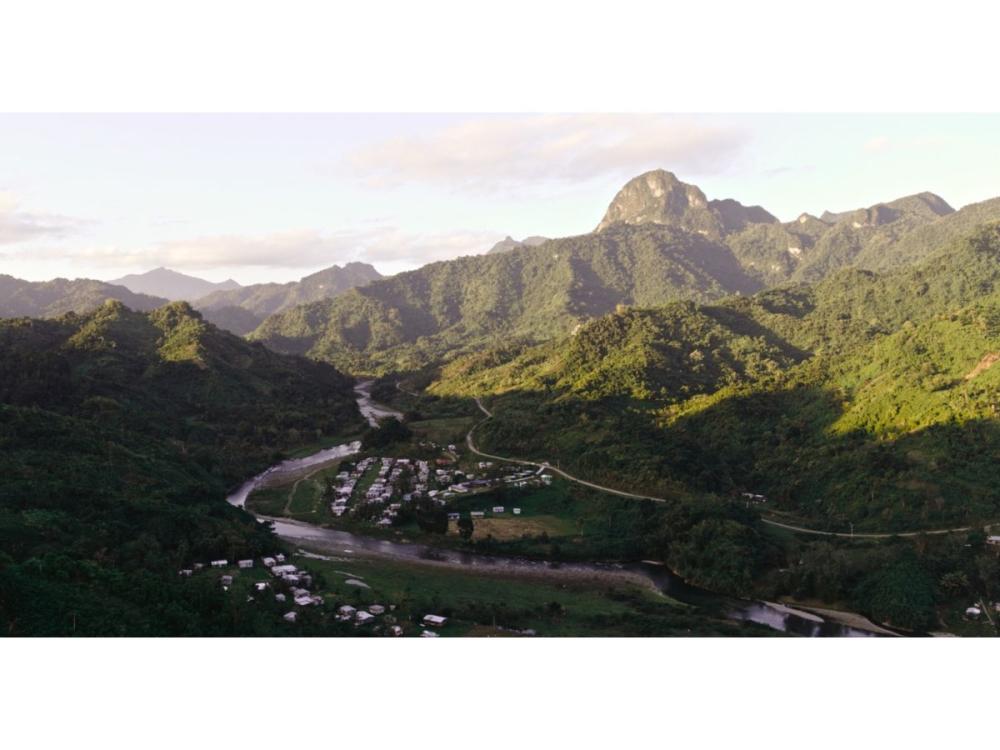 A view of green mountains with a river winding through them. Houses are gathered either side of the river.