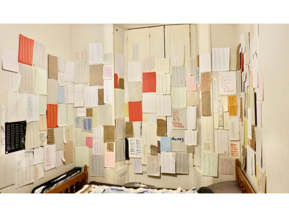 A studio, the walls completely covered in sheets of paper of different sizes and colours. They're all covered in writing, and the ones we can make out all have the word 'Thomas' written multiple times across the page