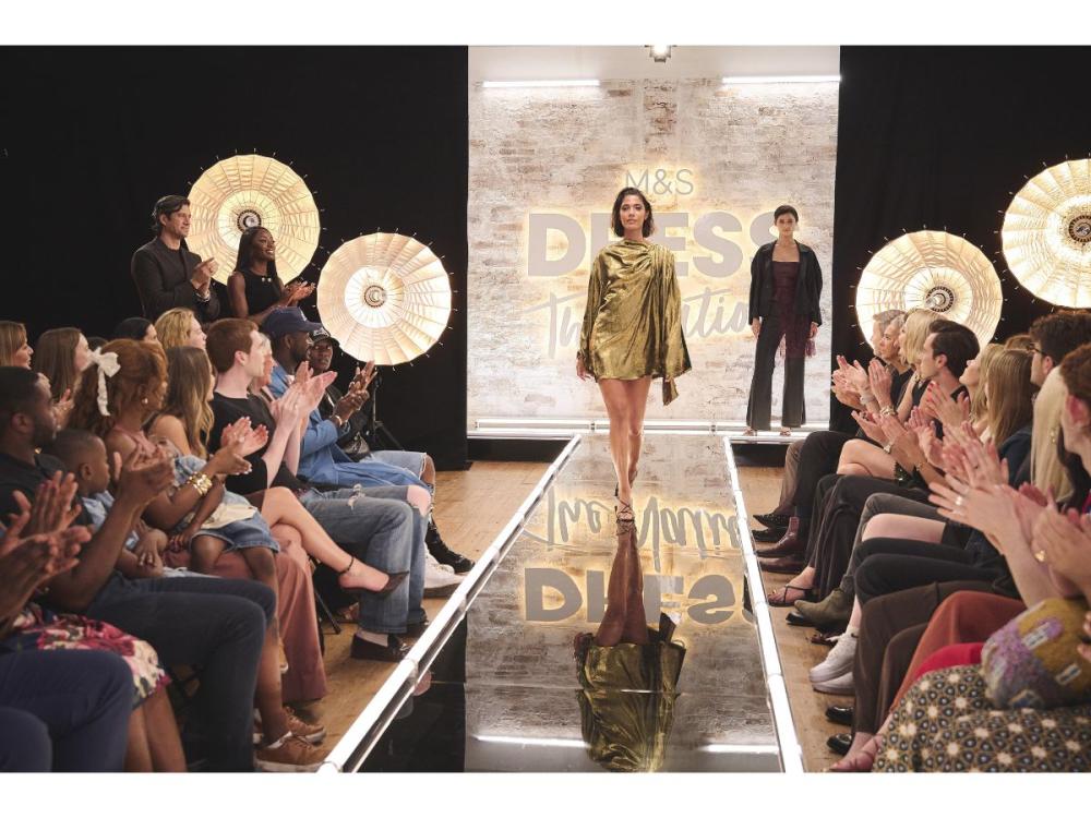 A model walks down a reflective catwalk, wearing the gold mini dress. The audience, lining the catwalk, applaud. Presenters AJ Odudu and Vernon Kay stand at the back in front of large lights, also applauding.