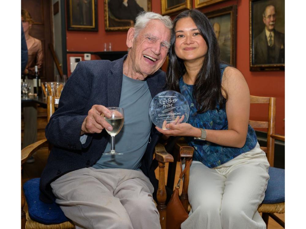 Emma sits with Klaus Flugge. They are both smiling and each has an arm around the other. She holds her award, he holds a glass of wine.