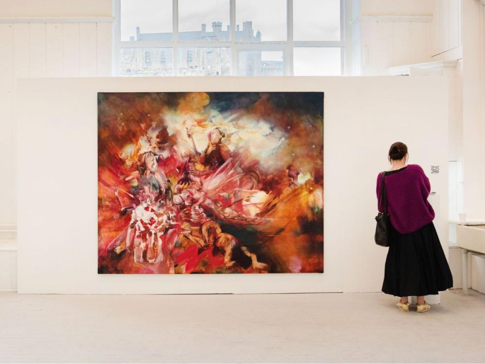 A large painting hung on a white wall; it is abstract - an explosion of energy and light with human figures included. A woman stands to the right of the painting; Through a window above, Edinburgh Castle is visible.