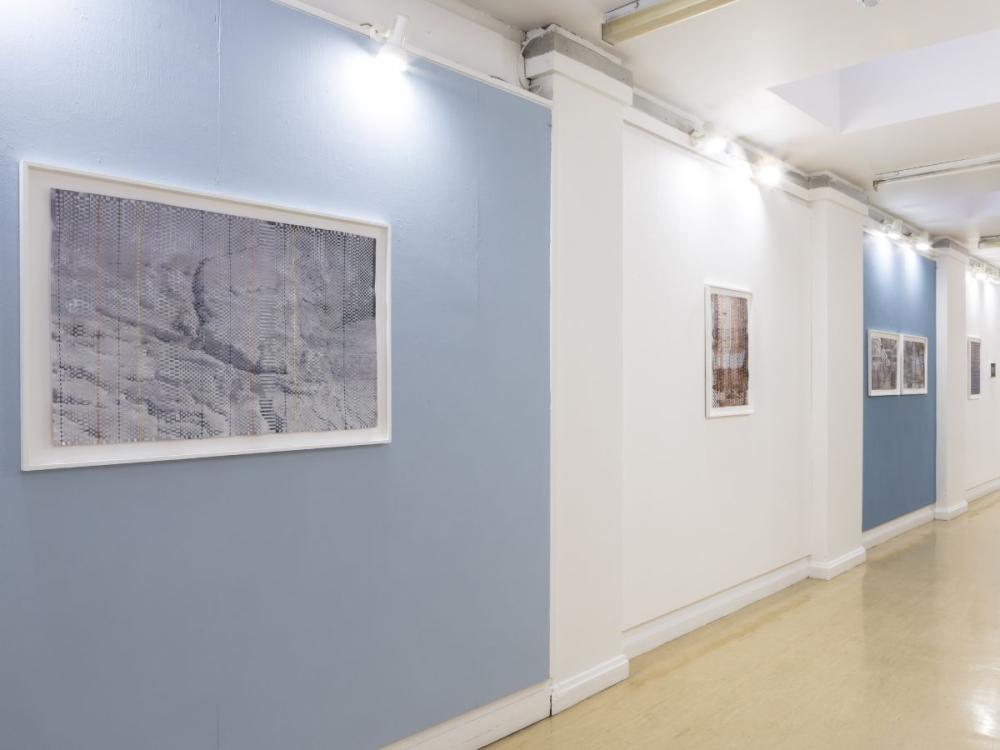 A series of paintings that are intricately woven from strips of paper, hung on blue and white walls; the nearest is pale blue, white and grey in colour.