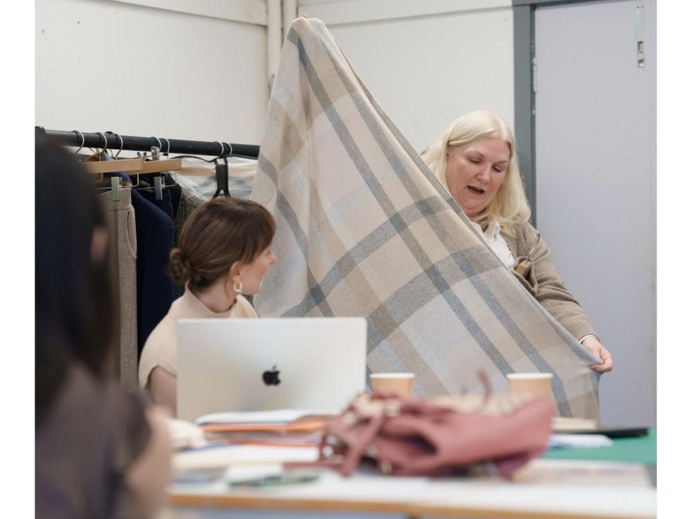 A woman, standing holds a piece of cream, beige and grey checked fabric between her outstretched arms. Another woman, seated, looks on.