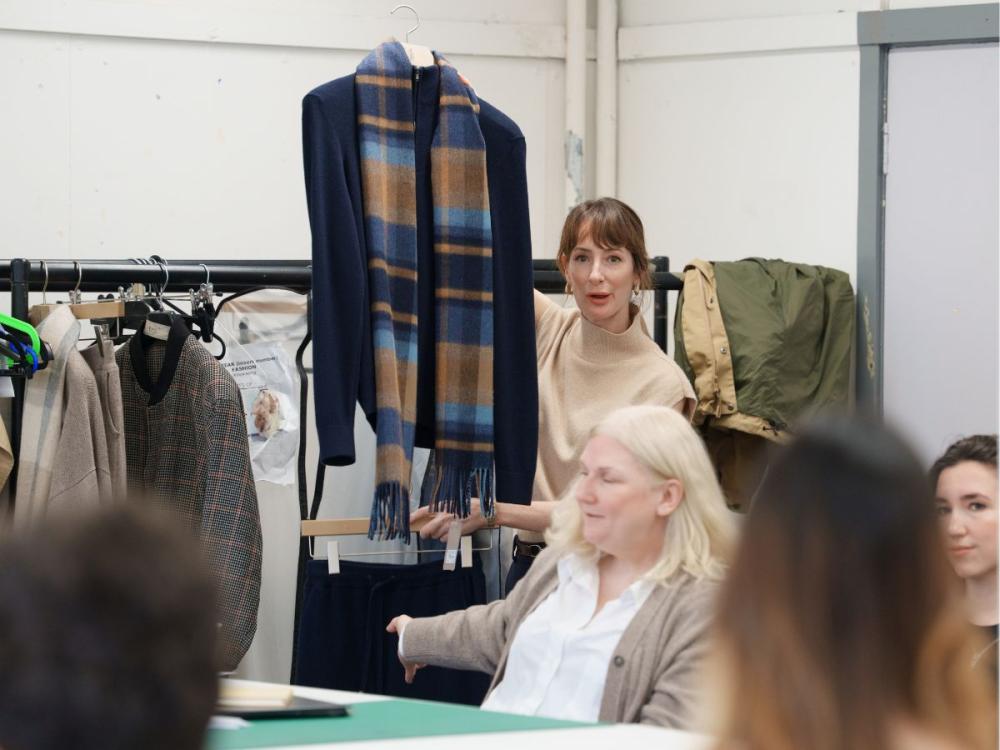 A woman, standing holds up a dark coloured sweater with a tartan scarf, while talking to seated students.