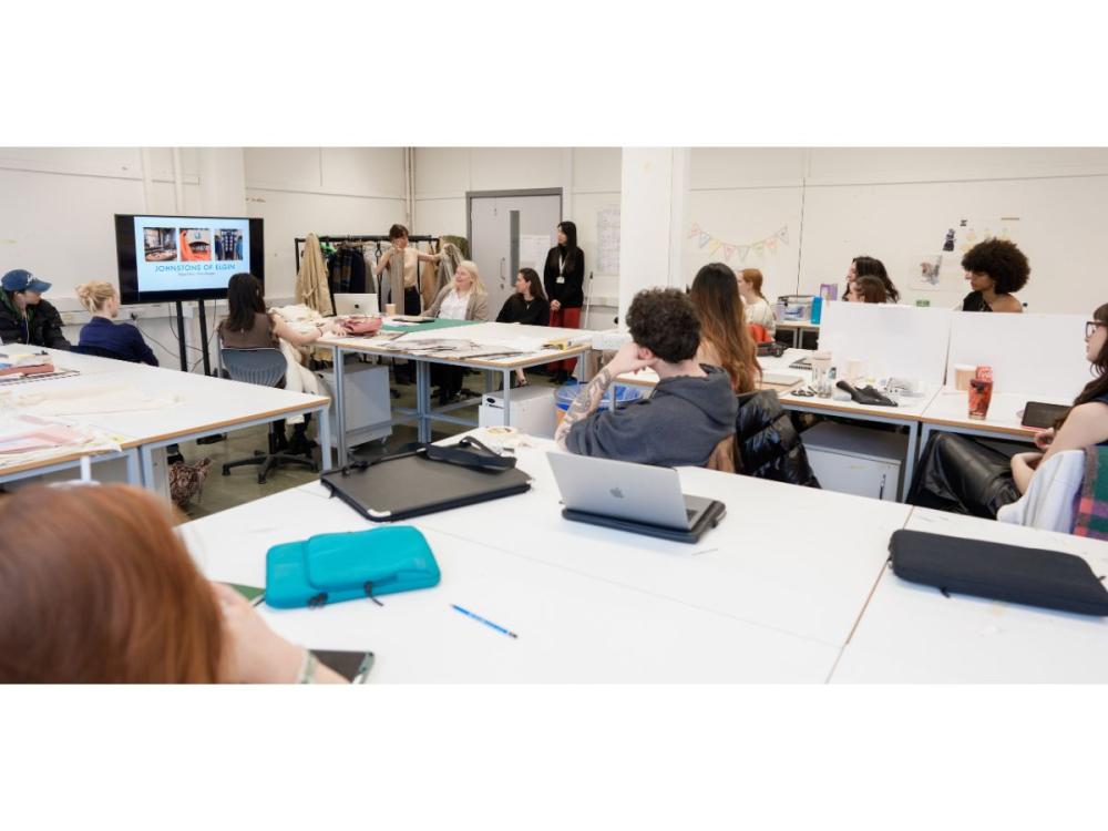 A room full of students, seated at desks, listens to a woman who is presenting at the front, holding two pieces of clothing aloft. A TV screen shows three photographs and the words Johnstons of Elgin.