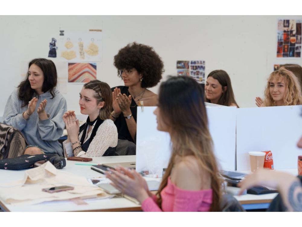 A group of students applauding. They're seated at desks strewn with paper, laptops, phones, etc. There are some drawings pinned to the wall behind them.