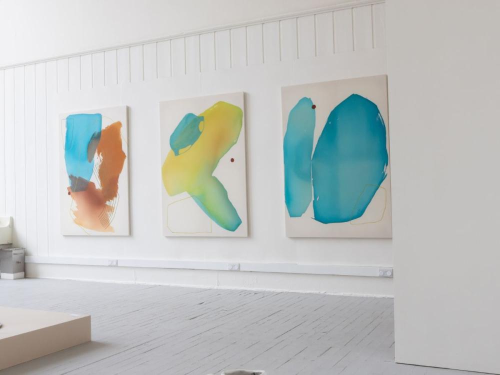 Large, abstract paintings hung on white gallery walls. Each shows huge pools of pale colour - pale blues, yellow and rust shades.