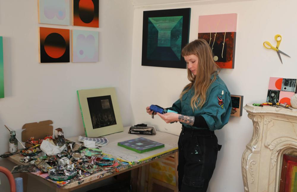 Artist Charlotte Roseberry is standing looking at her work on the BA bag. She is in her studio and there are artworks and materials around her.