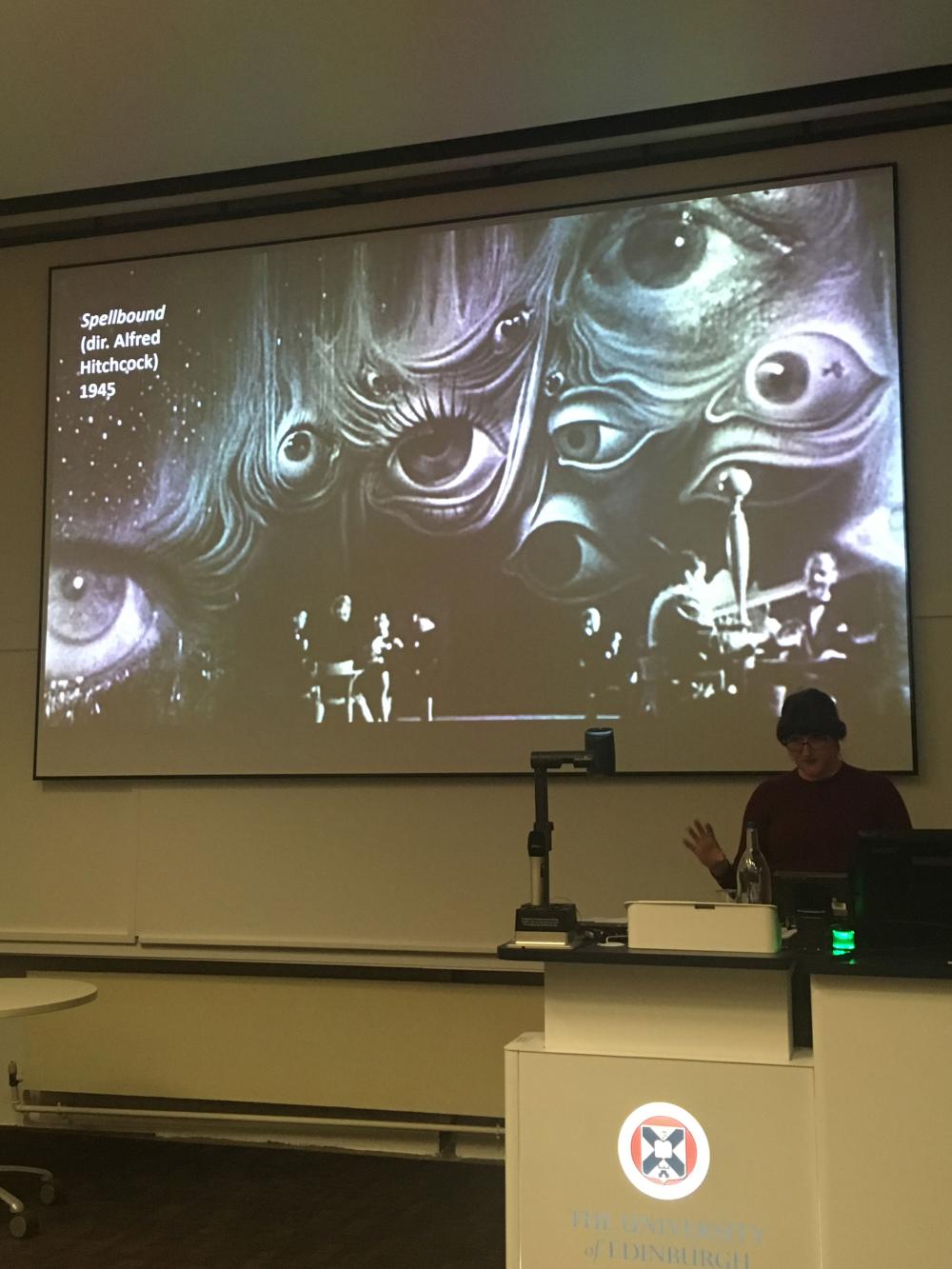 A person stands at a desk giving a presentation on a large screen. An image of a scene from Alfred Hitchcock's film 'Spellbound' featuring people sitting at tables whilst many different sized eyes hover above them.