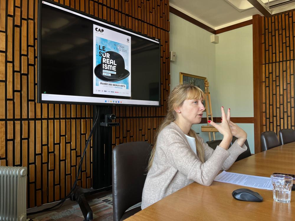 A person sitting in the wooden paneled Boardroom at ECA, giving a presentation in front of a screen with a picture of a bowler hat on it and text that reads 'Le Surrealisme'