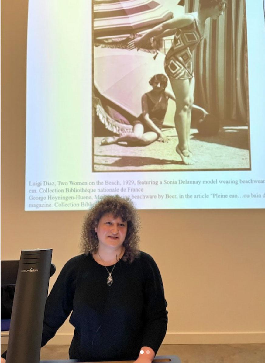 A person delivers a lecture in front of a projected image of a woman in a vintage 1930s swimsuit, standing on her toes whilst another woman is sitting nearby and smiling at the camera.