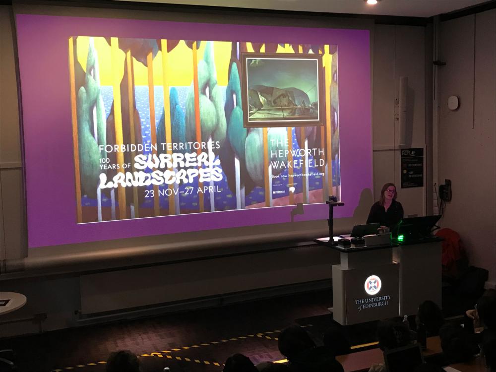 A person delivers a lecture in front of a screen with a purple graphic on it and surrealist landscape images.