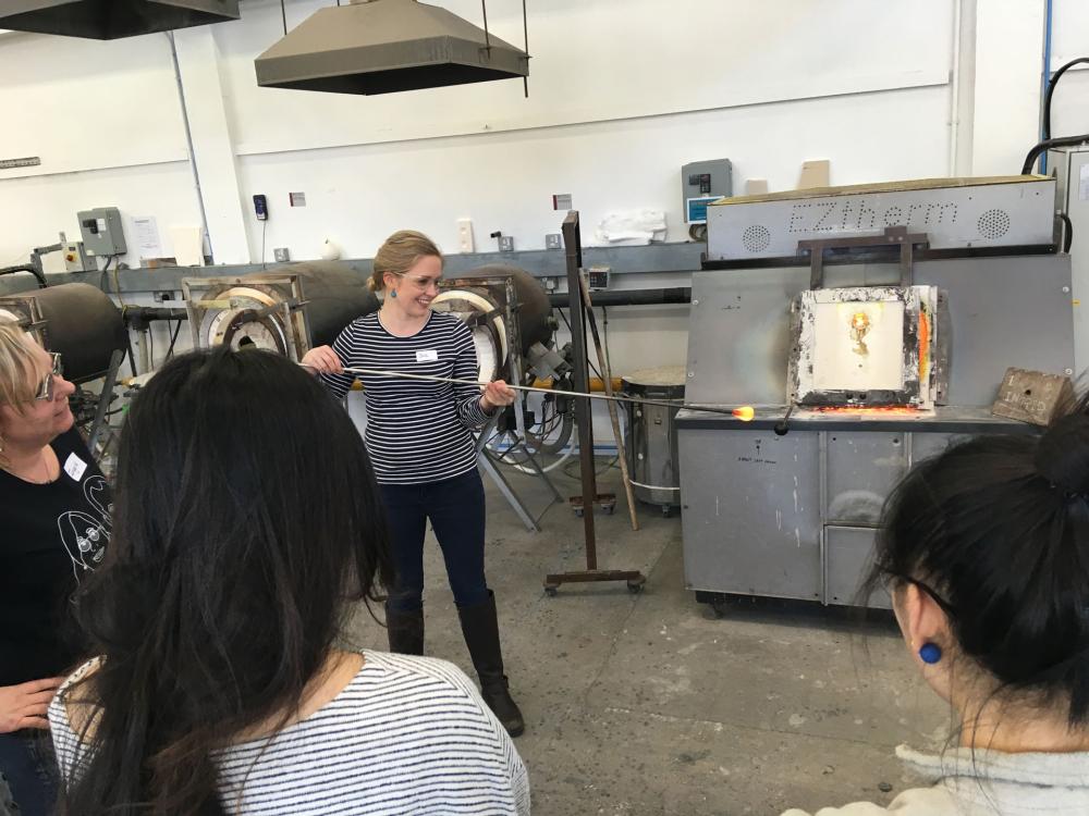 Head of Design, Dr Jessamy Kelly holds a long pole with a hot bulb of glass on the end of it. Students are gathered around for the demonstration.