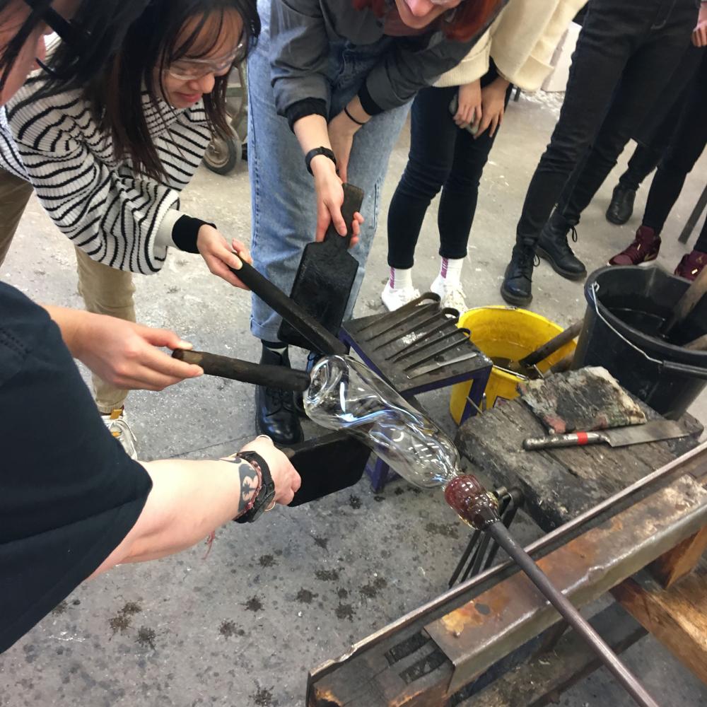 A group of students take part in a surrealist glass blowing workshop, shaping a piece of hot glass.