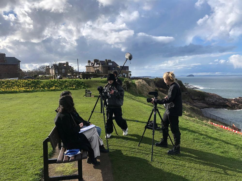 Camera crew outdoors filming people on the bench. Sea, park and buildings in the background.