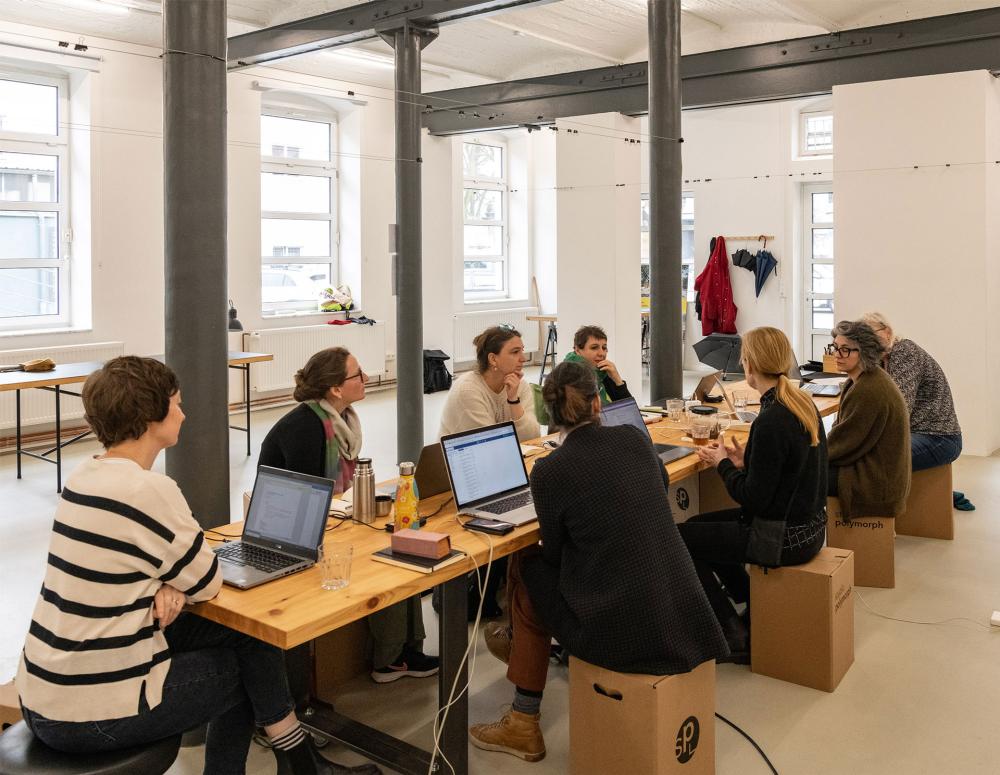A group of people with laptops sitting around a long wooden table, having an editorial meeting.