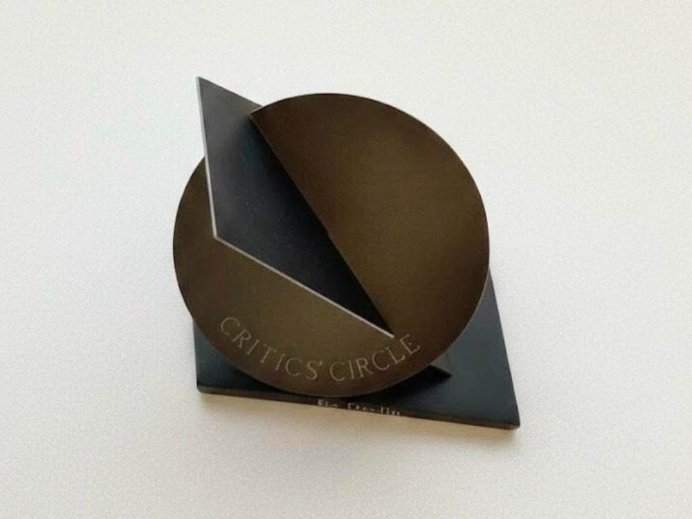 The award, comprising a circle of dark-coloured metal with the words 'Critics' Choice' in caps at the bottom; There's an intersecting, angular piece of metal and another forms the base, both darker than the circle