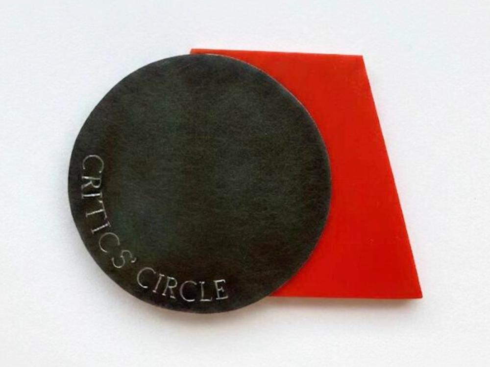 The award, comprising a dark-coloured circle of metal with the words 'Critics' Circle' in caps on the bottom part, and behind it a red-coloured trapezoid.