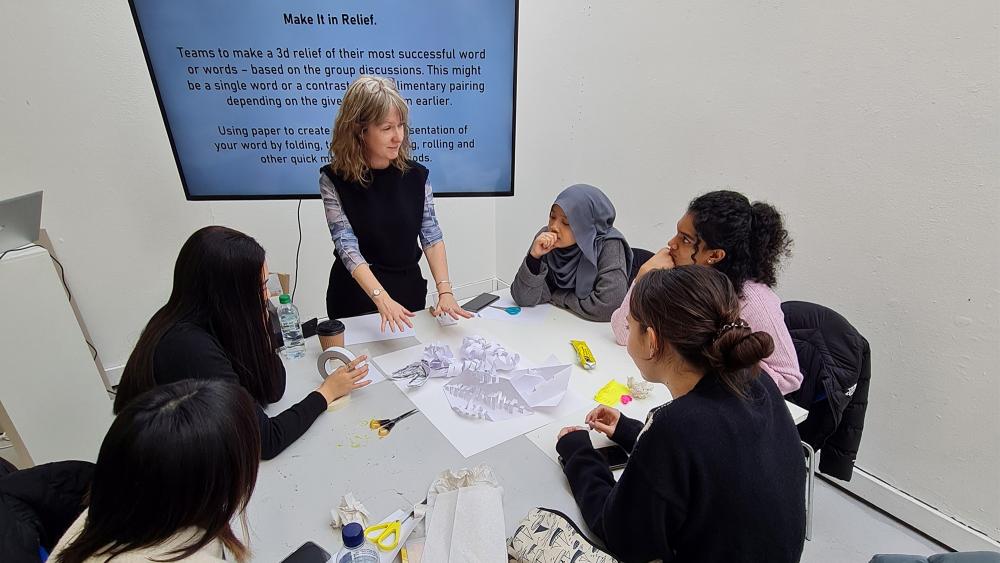 a group of interior design students around a table listening to a female teacher with a powerpoint presentation.