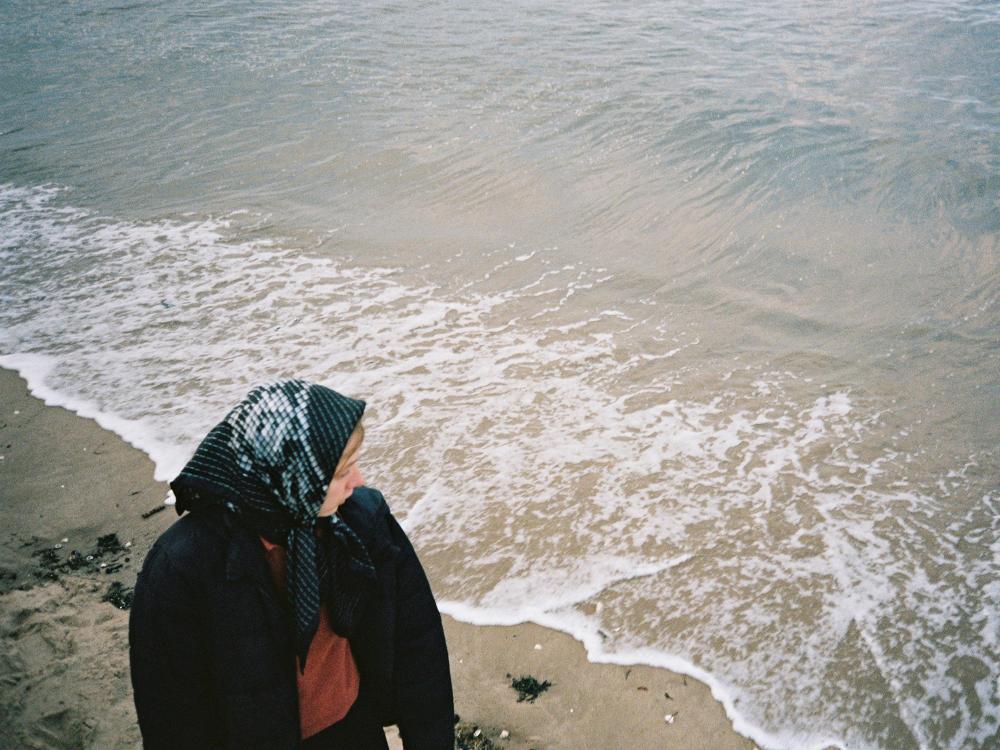 Woman walking on the waters edge at the beach, looking out to sea. She wears a black printed silk scarf. 